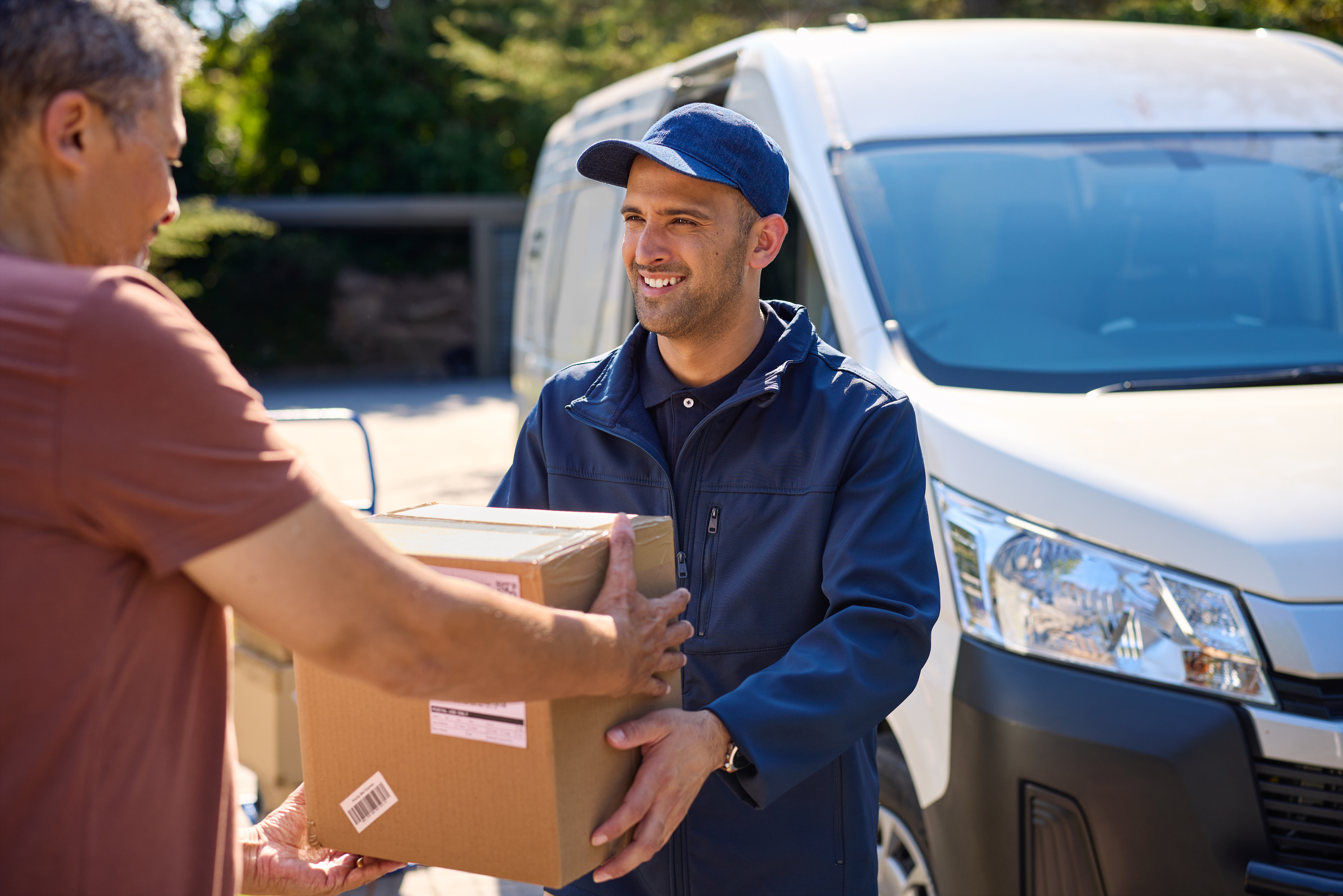 Smiling delivery man in blue uniform delivering parcel box to a mature man. Happy postal man delivering parcel to customer at home. Multiethnic elderly man receiving package from courier in the driveway outside the house.