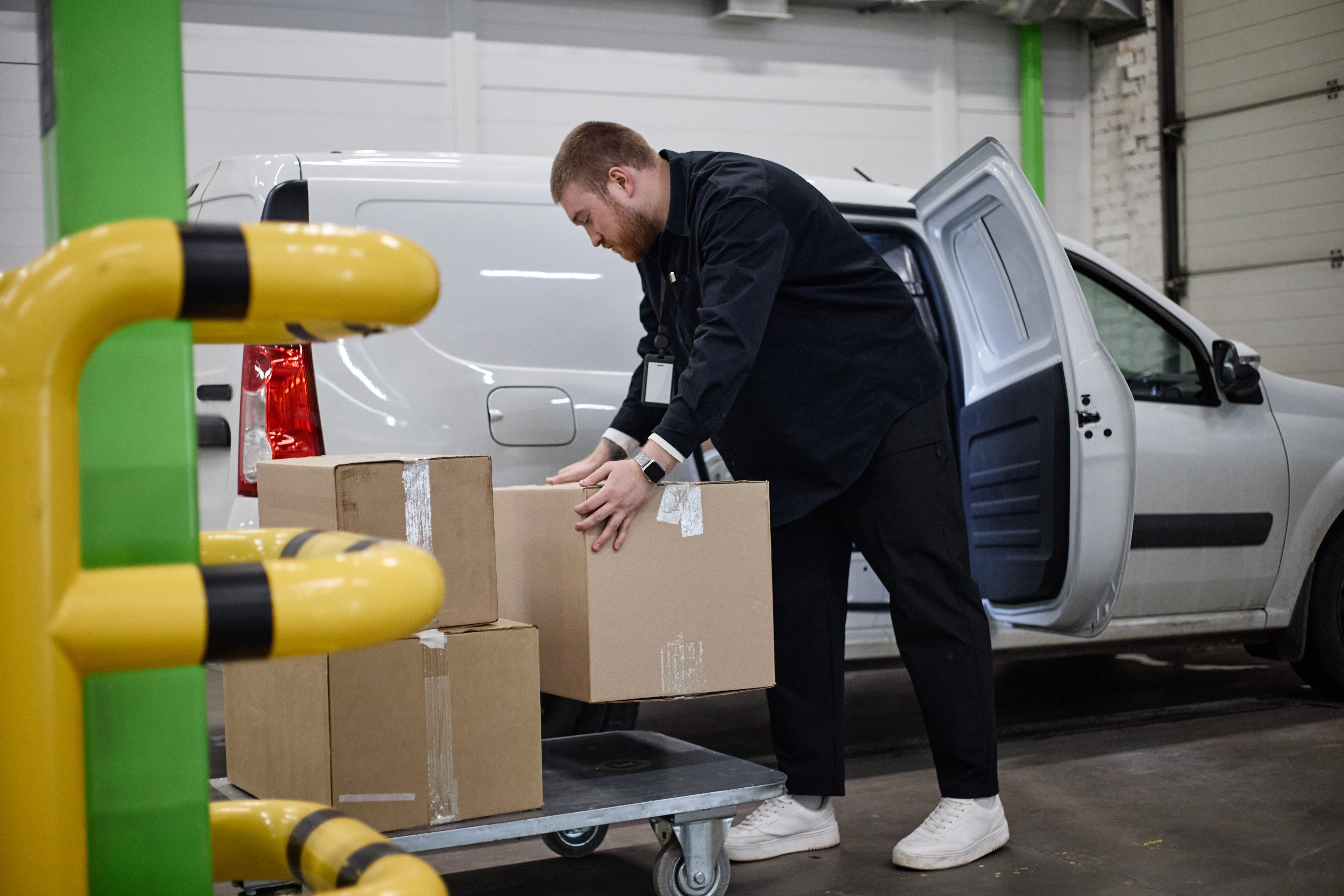Caucasian young adult delivery worker loading cardboard boxes beside cargo van in storage facility, handling parcel logistics. Useful for shipping, warehousing, fulfillment marketing